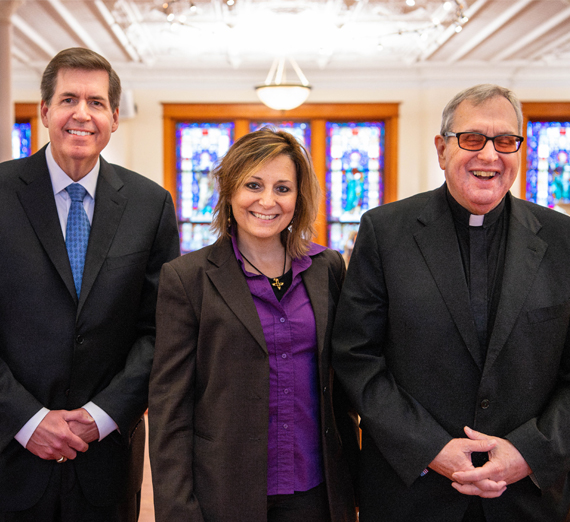 A group of five people stand together in a brightly lit hospital hallway, smiling at the camera. Two wear stethoscopes and medical attire, one wears a Gonzaga University sweatshirt, and others are dressed in casual or professional clothing, representing a mix of students and healthcare professionals.
